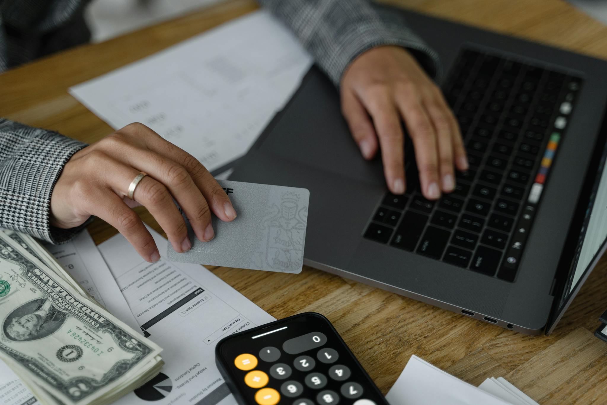 Close-up of a person using a credit card and laptop in a financial setting with cash and calculator.